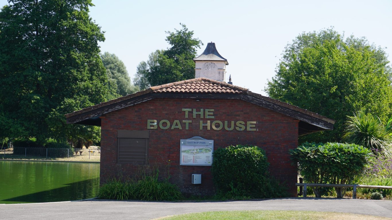 A red-brick boathouse with a sign, surrounded by greenery and water.