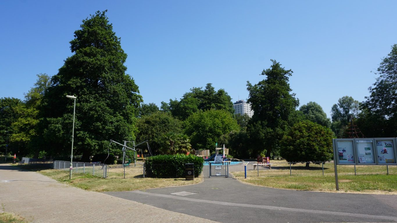 A sunny park with trees, grass, and a pathway leading to a fenced playground.